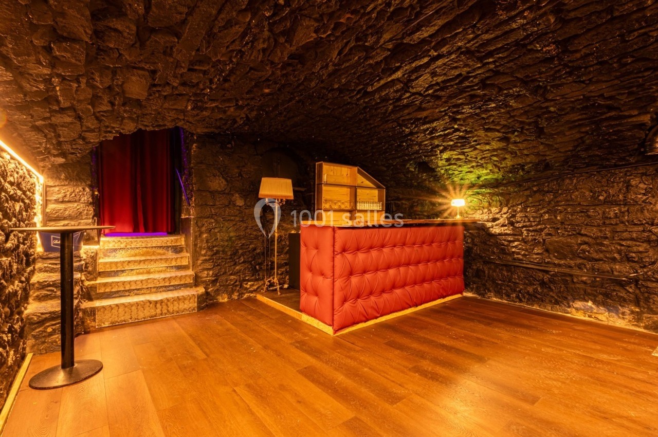 Salle voûtée en pierre avec un comptoir rouge, éclairage chaleureux et escalier menant à un rideau rouge.