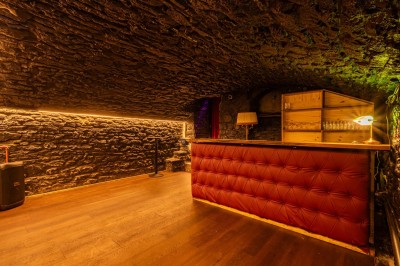 Miniature Speakeasy privatisable Salle voûtée en pierre avec un comptoir rouge, éclairage chaleureux et escalier menant à un rideau rouge.
