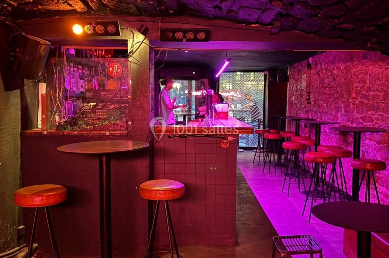 Bar à l'ambiance tamisée avec éclairage violet, comptoir central, tabourets hauts et murs en pierre apparente.