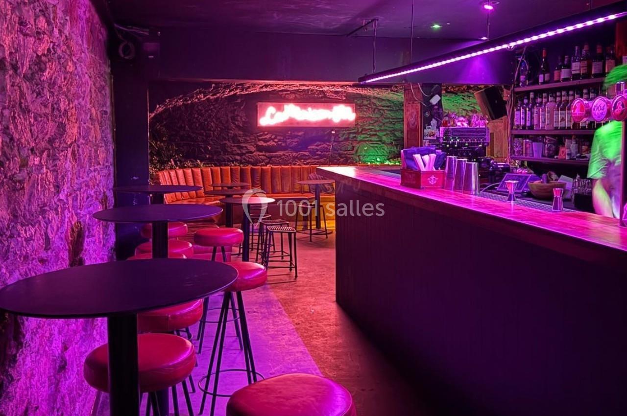 Bar à l'éclairage violet avec comptoir en bois, tabourets rouges et banquettes en fond près d'un mur en pierre.