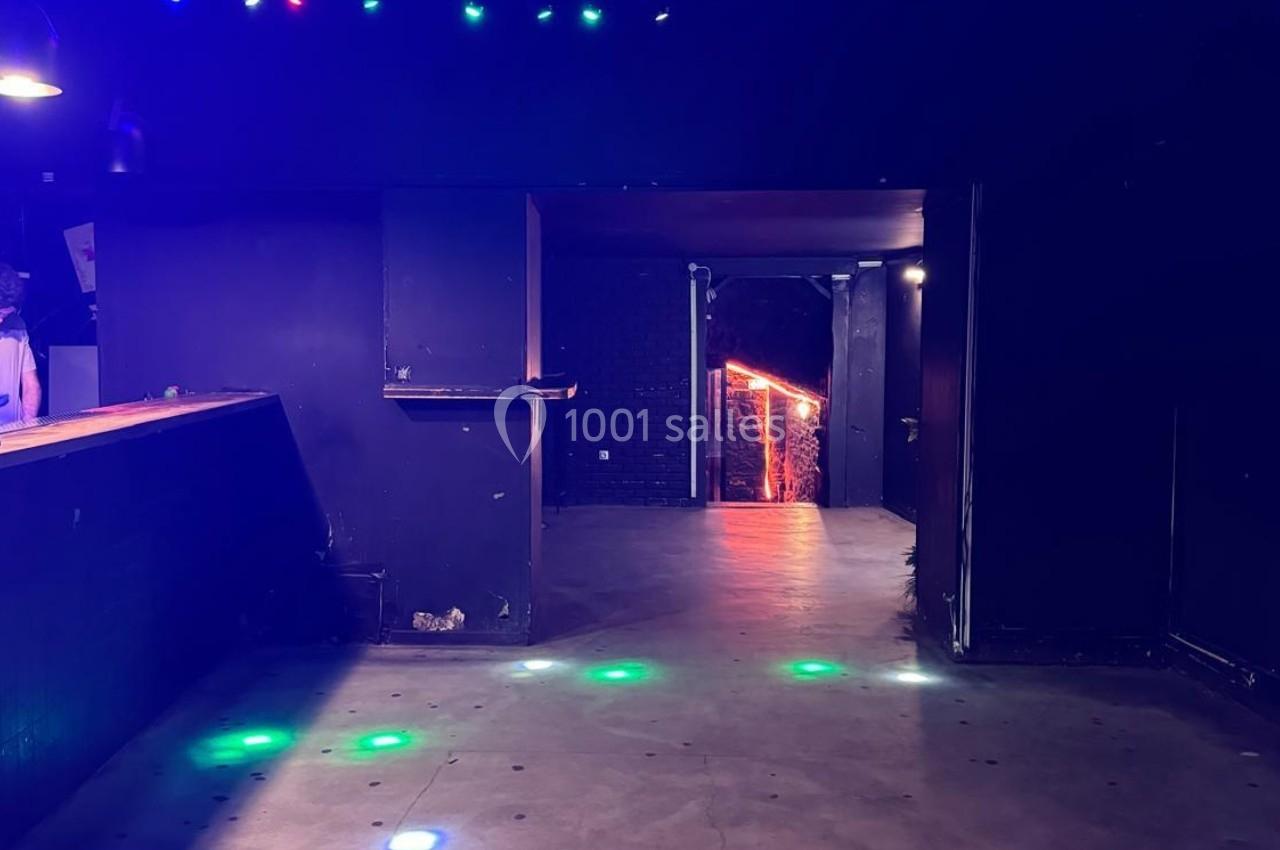 Salle sombre avec un comptoir à gauche, sol en béton et lumières colorées projetées au sol.