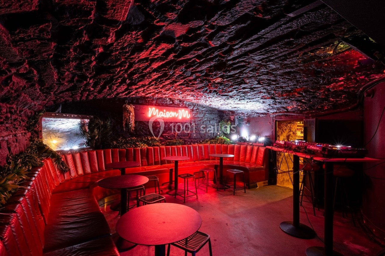 Salle voûtée avec banquettes rouges, tables basses, éclairage néon rose et espace DJ à droite.
