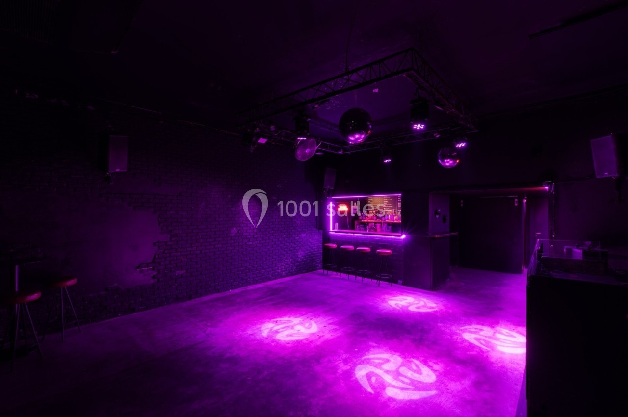 Salle sombre avec éclairage violet, piste de danse vide, bar éclairé au fond et murs en briques noires.