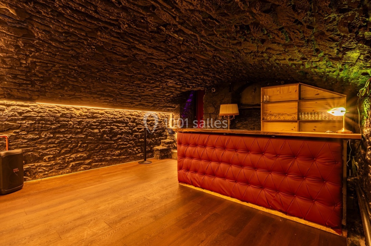Bar en bois avec comptoir capitonné rouge dans une cave voûtée en pierre, éclairée par une lumière chaude.