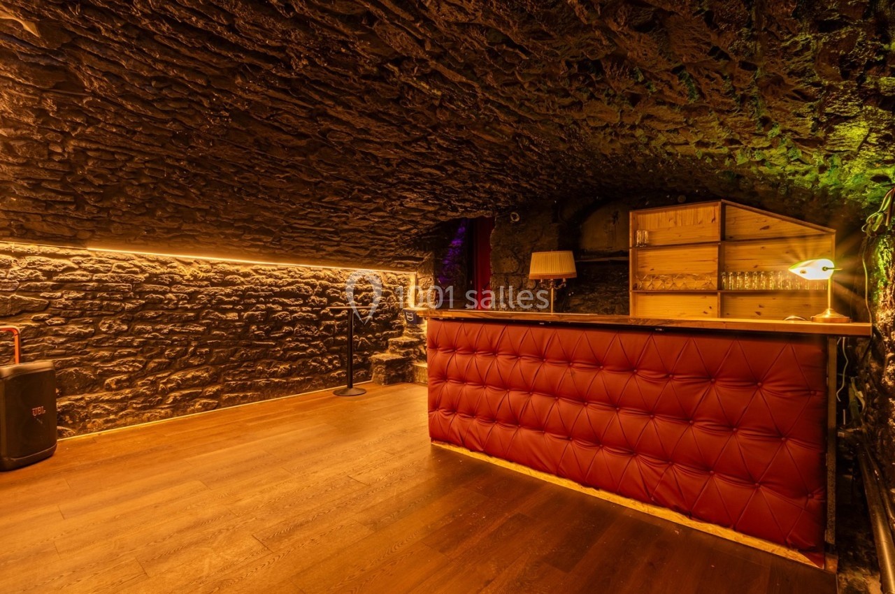 Cave voûtée en pierre avec un bar en bois et cuir rouge, éclairage chaleureux et étagères avec verres.