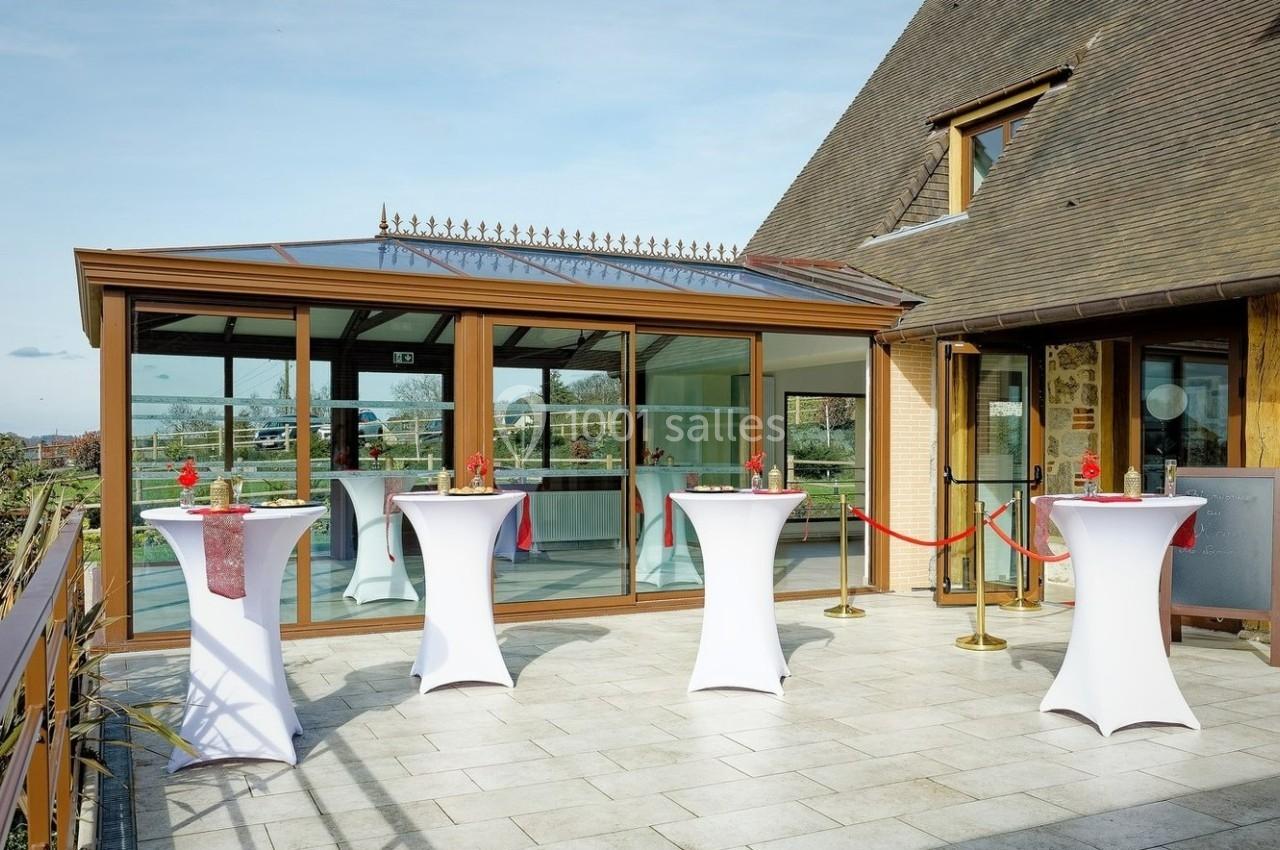 Location salle Saint-Samson-de-la-Roque (Eure) - Manoir Néo Normand #4 Terrasse avec tables hautes habillées de nappes blanches, devant une véranda vitrée et un bâtiment en bois.