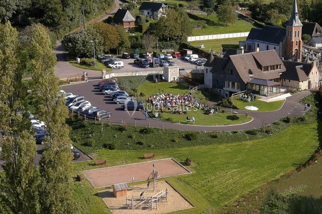 Location salle Saint-Samson-de-la-Roque (Eure) - Manoir Néo Normand #12 Vue aérienne d'un village avec une église, un parking, une aire de jeux et un rassemblement de personnes en extérieur.