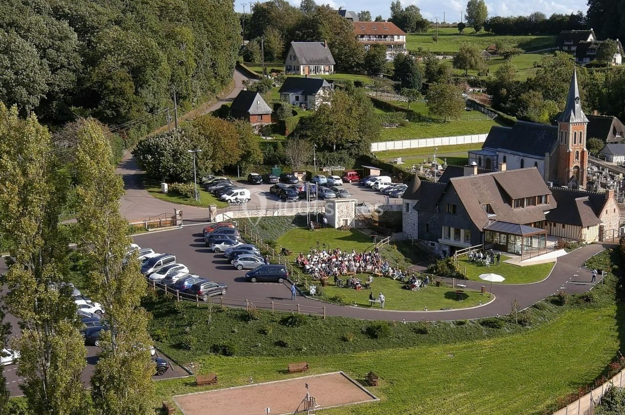 Location salle Saint-Samson-de-la-Roque (Eure) - Manoir Néo Normand #15 Vue aérienne d'un village avec une église, un parking, une aire de jeux et des personnes rassemblées sur une pelouse.