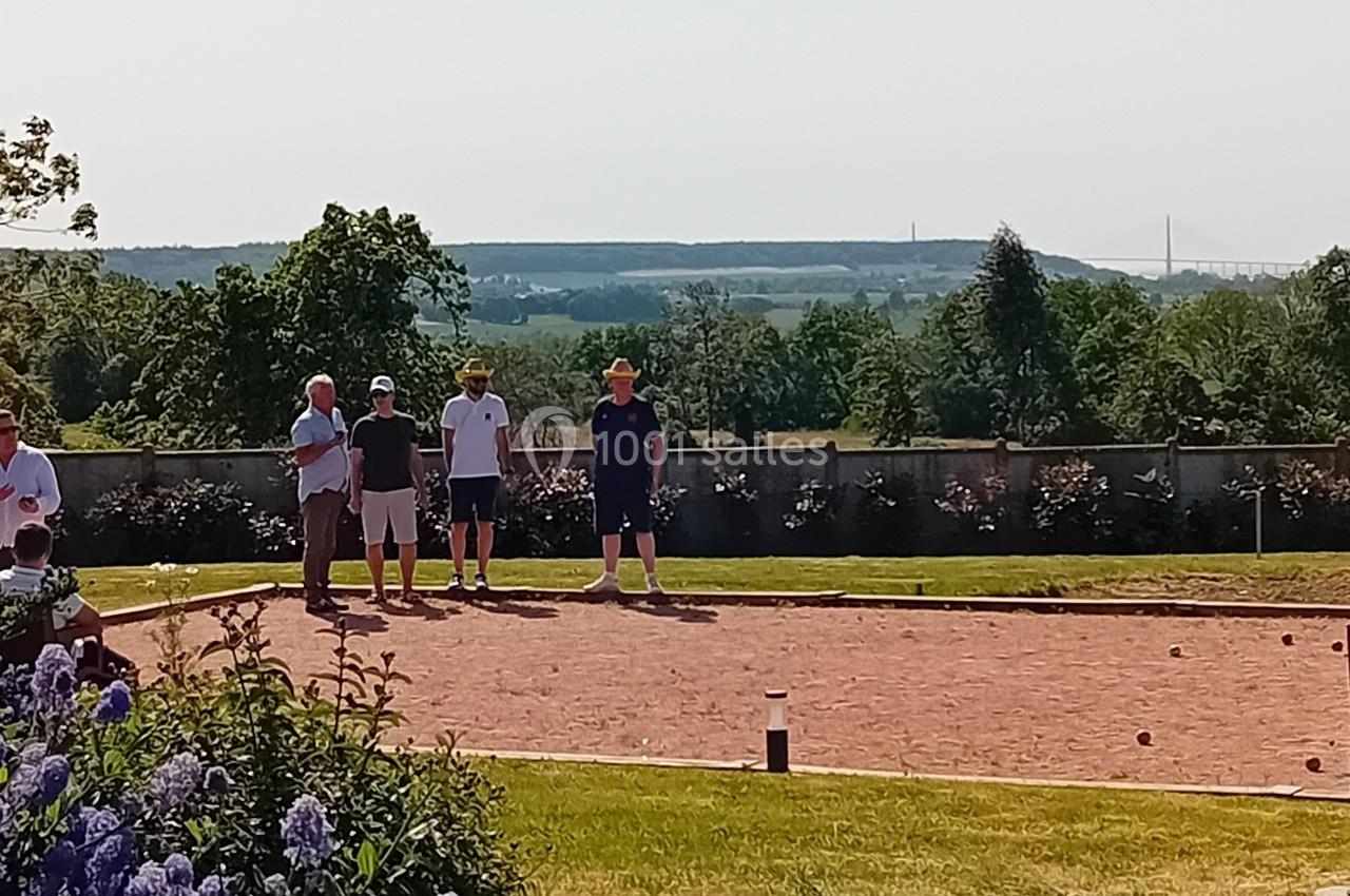Location salle Saint-Samson-de-la-Roque (Eure) - Manoir Néo Normand #18 Des personnes jouent à la pétanque sur un terrain ensoleillé, avec un paysage verdoyant et un pont visible au loin.