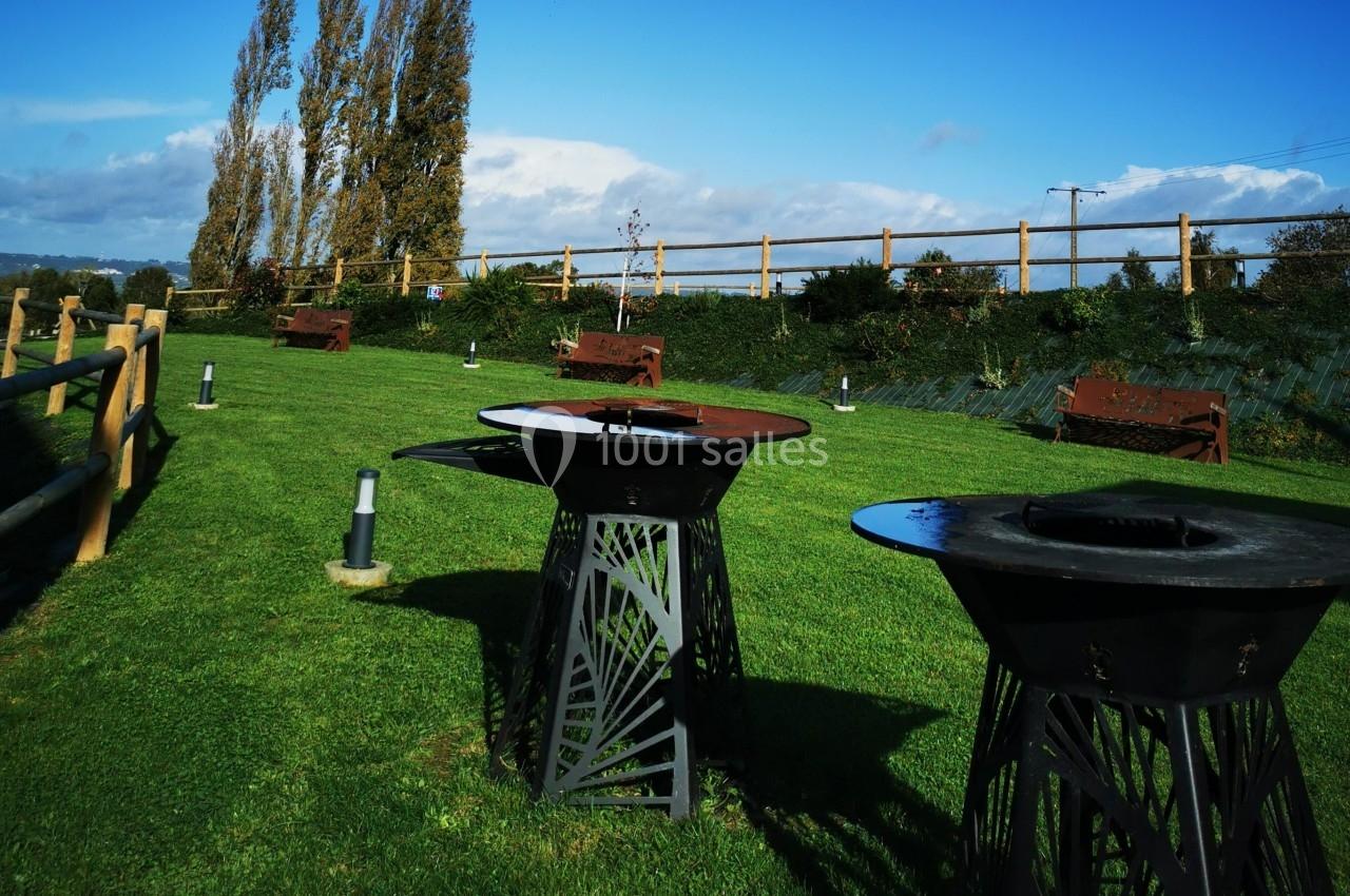 Location salle Saint-Samson-de-la-Roque (Eure) - Manoir Néo Normand #20 Barbecues en métal sur une pelouse verte, entourés de bancs en bois et d'une clôture en bois sous un ciel dégagé.