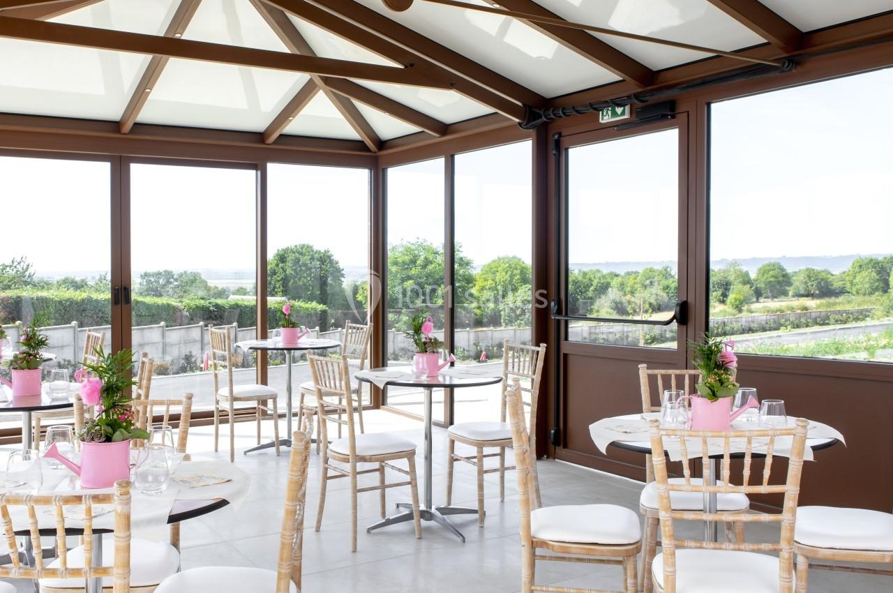 Location salle Saint-Samson-de-la-Roque (Eure) - Manoir Néo Normand #26 Salle lumineuse avec baies vitrées, tables rondes décorées de fleurs roses et chaises en bois, vue sur un paysage verdoyant.