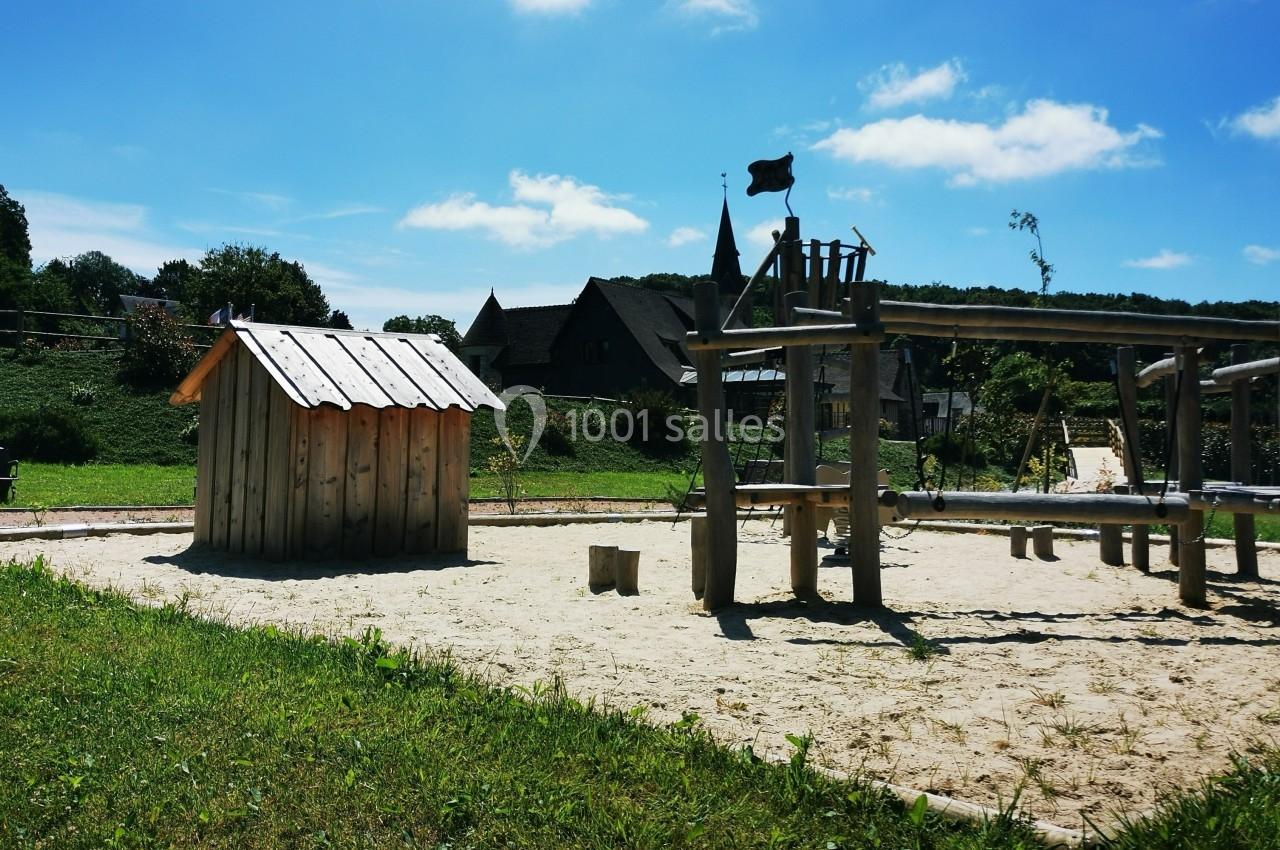 Location salle Saint-Samson-de-la-Roque (Eure) - Manoir Néo Normand #38 Aire de jeux en bois sur sol sablonneux, entourée de verdure et située près de bâtiments ruraux sous un ciel bleu.