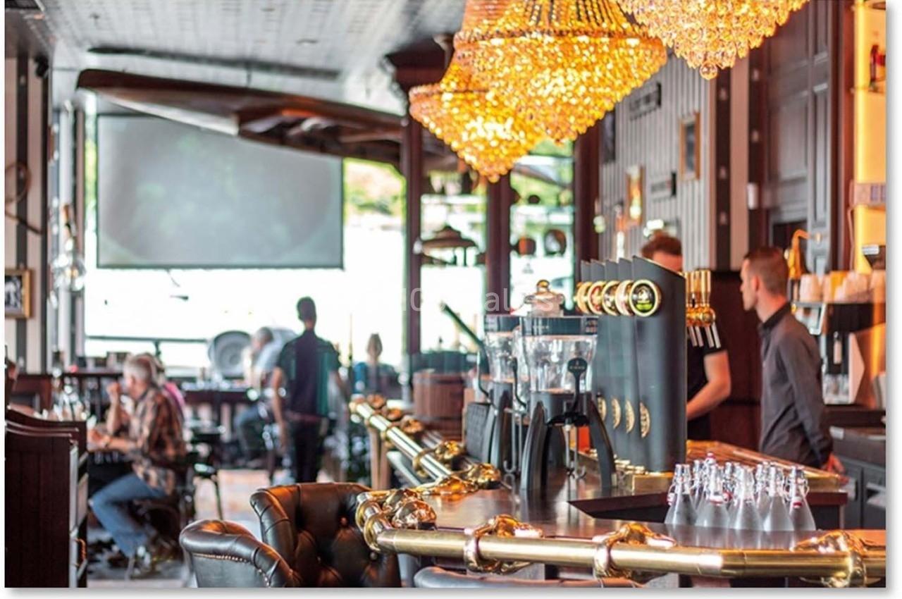 Intérieur d'un bar lumineux avec comptoir en bois, luminaires suspendus et clients assis ou debout.