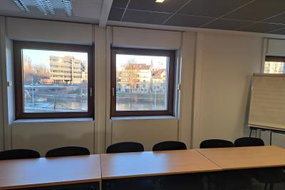 Salle de réunion avec des tables et des chaises alignées, vue sur des bâtiments et un cours d'eau à travers les fenêtres.