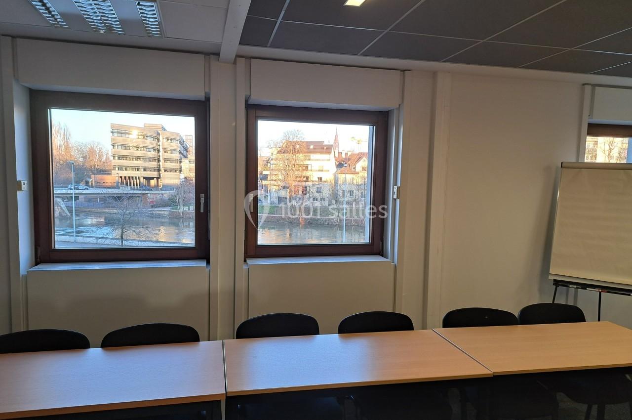 Salle de réunion avec des tables et des chaises alignées, vue sur des bâtiments et un cours d'eau à travers les fenêtres.