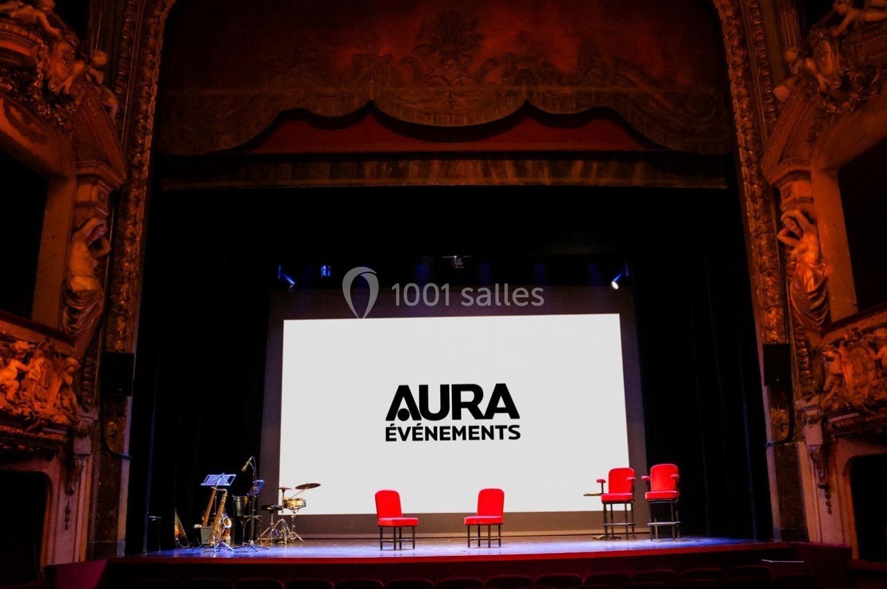 Scène de théâtre avec écran affichant ’Aura Événements’, quatre fauteuils rouges et des instruments de musique.