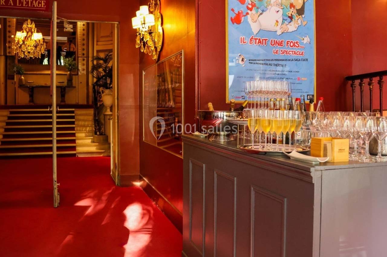 Buffet avec verres de champagne dans un hall rouge, escalier et affiche de spectacle en arrière-plan.