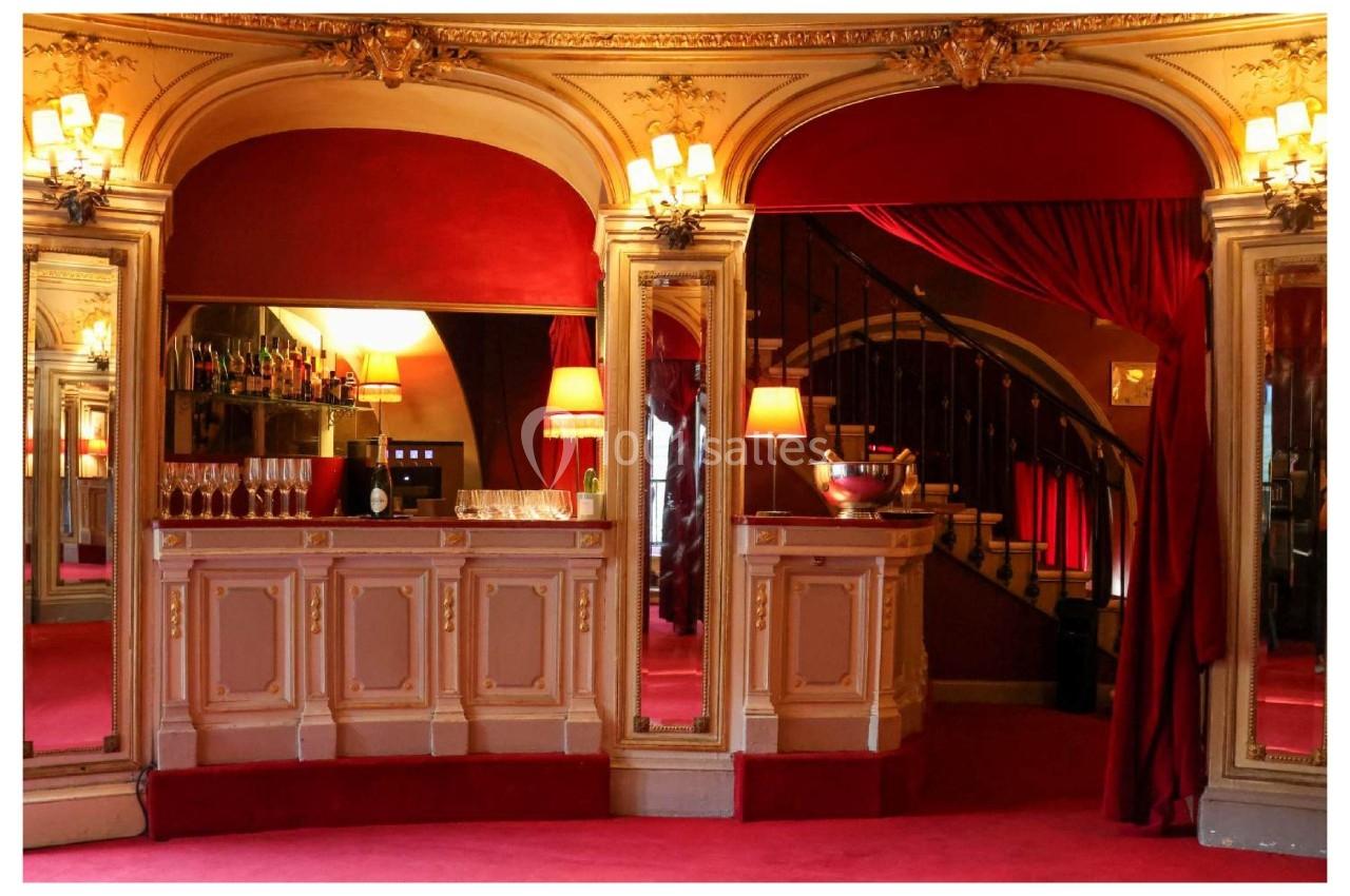 Bar élégant avec comptoir en bois sculpté, miroirs, lumières tamisées et rideaux rouges dans un décor classique.