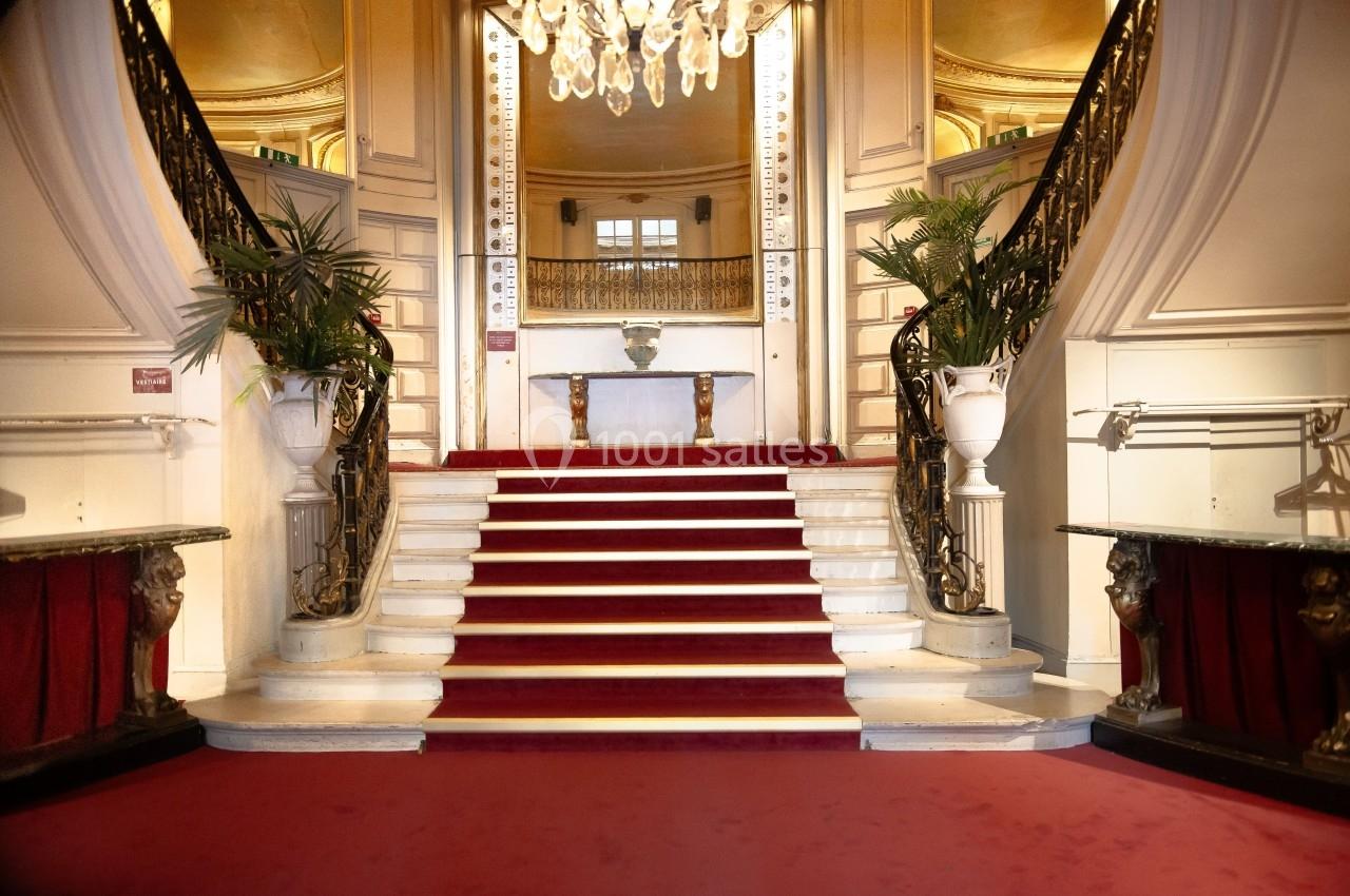Escalier central avec tapis rouge, entouré de rampes ornées et de plantes, dans un intérieur élégant et lumineux.