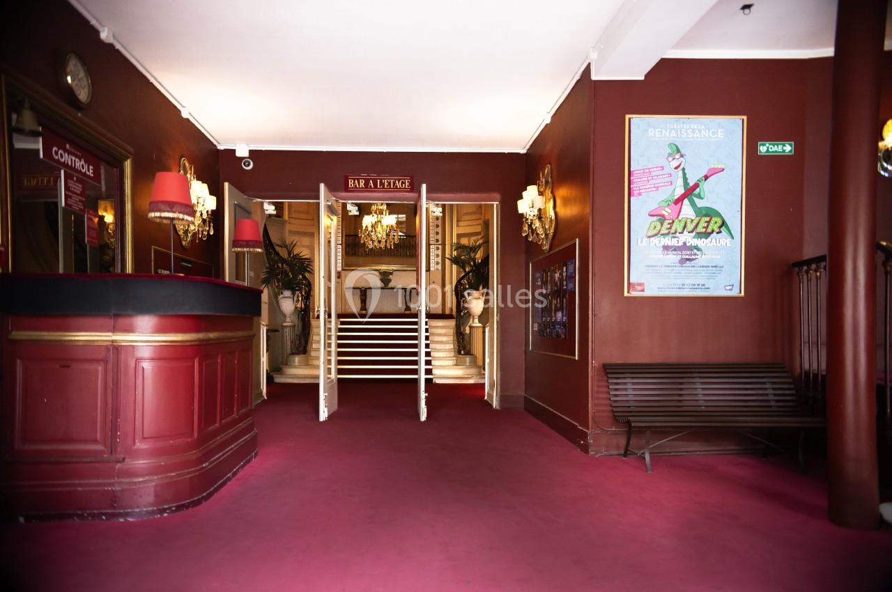 Entrée d'un théâtre avec comptoir rouge, escalier central, affiches murales et éclairage tamisé.