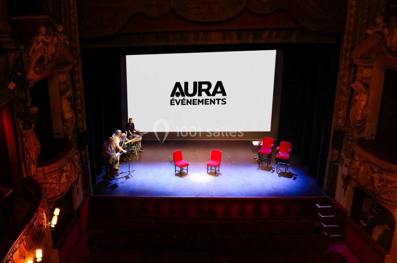 Scène d'un théâtre avec écran affichant ’Aura Événements’, deux chaises rouges et musiciens en répétition.