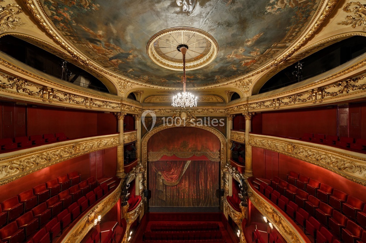 Intérieur d'un théâtre ancien avec balcon, plafond peint, lustre central et rideau de scène rouge.