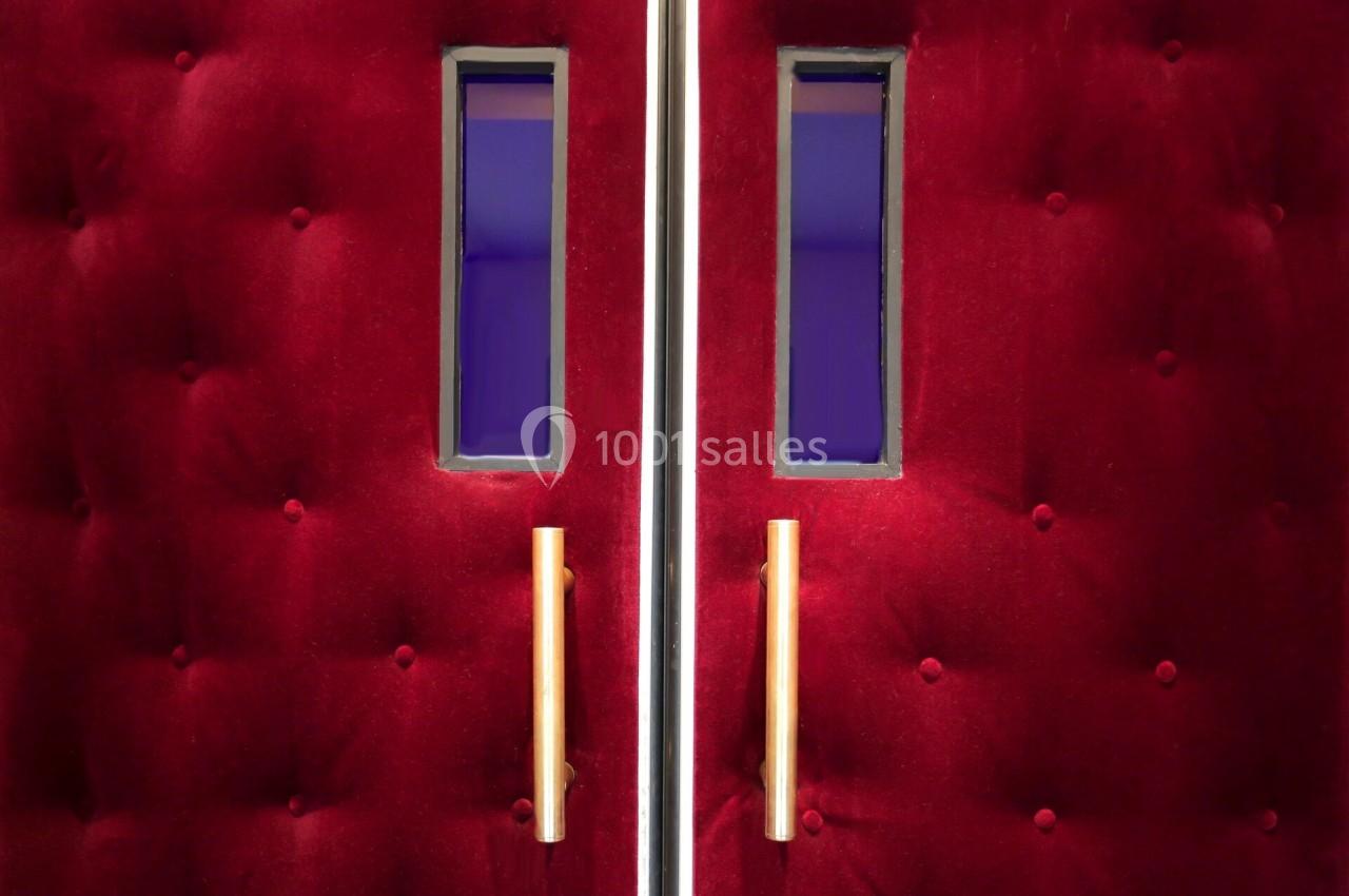 Portes capitonnées rouges avec poignées dorées et petites fenêtres rectangulaires laissant entrevoir une lumière violette.