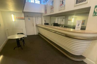 Comptoir rétro dans un hall avec une table et deux chaises, affiches murales et signalétique visible.