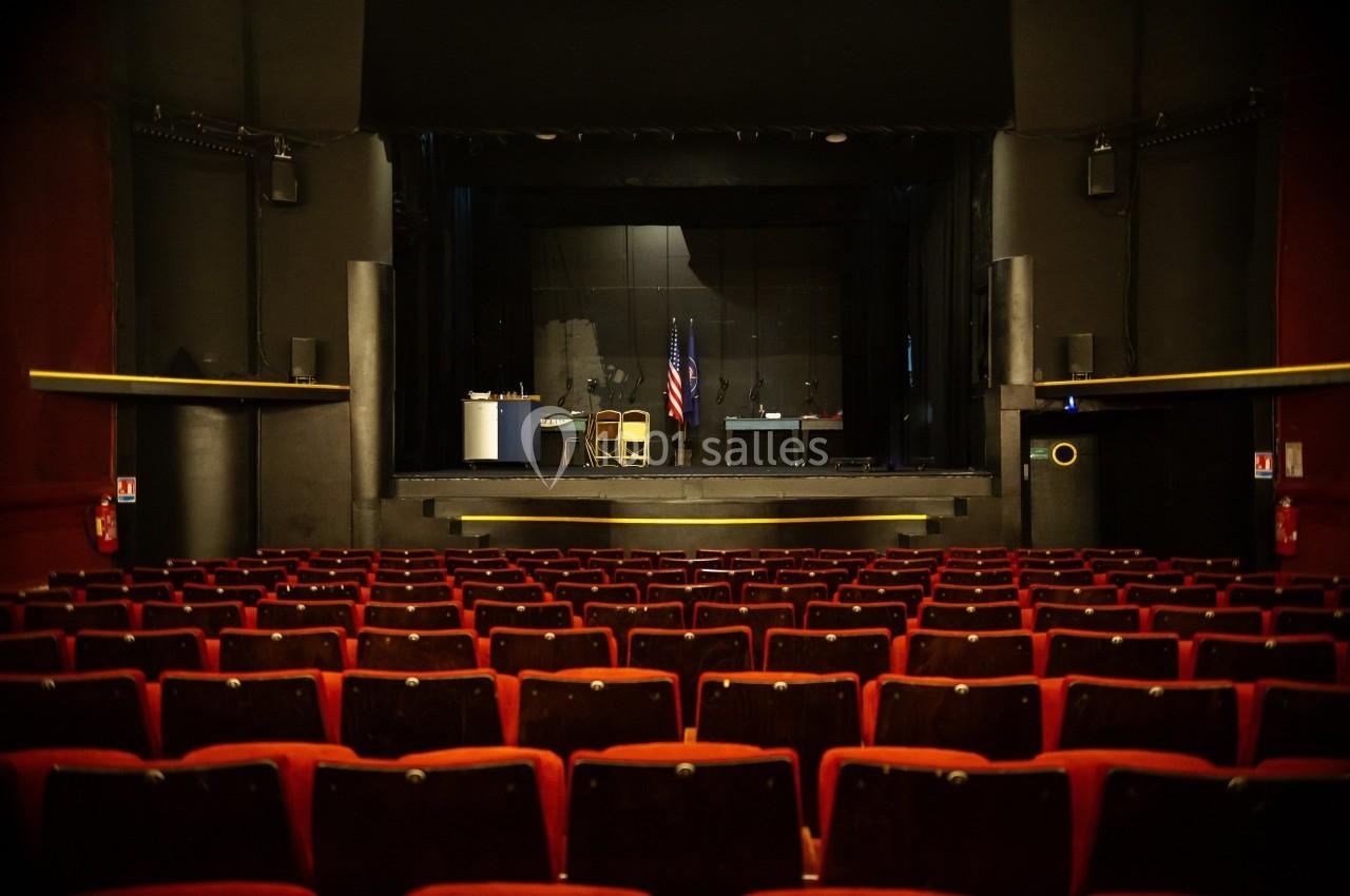 Salle de théâtre vide avec des rangées de sièges rouges et une scène équipée de drapeaux et de mobilier.