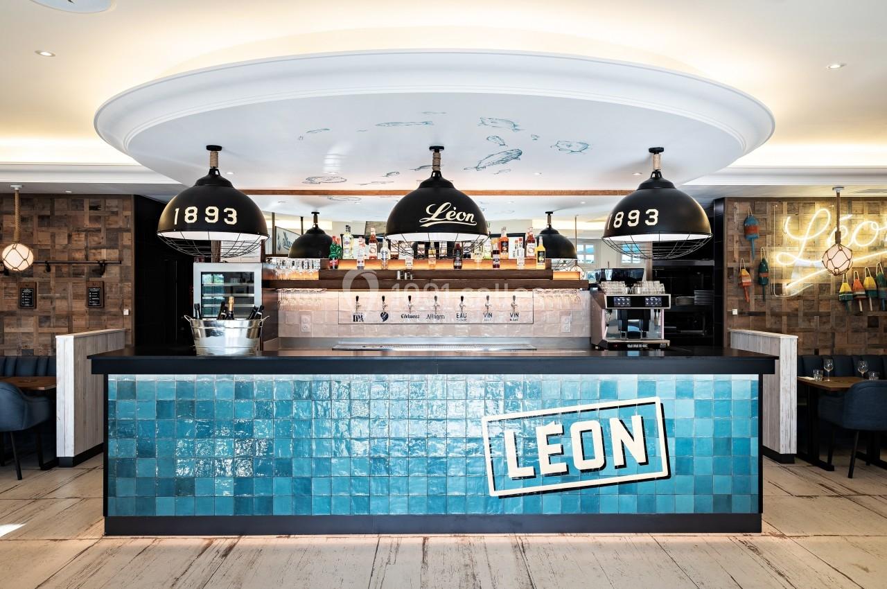 Comptoir de bar avec carrelage bleu, luminaires suspendus noirs et étagères garnies de bouteilles en arrière-plan.