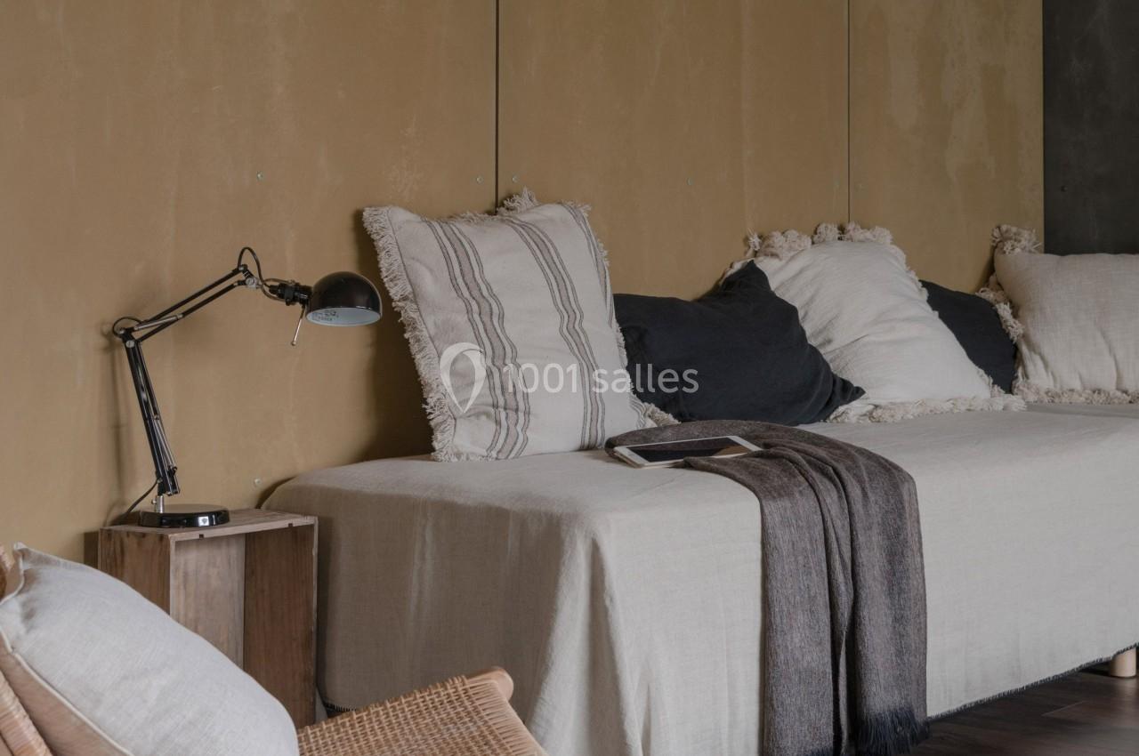 Salon minimaliste avec canapé beige, coussins texturés, plaid, lampe de lecture et fauteuil en bois sur parquet sombre.