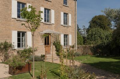Façade en pierre d'une maison à trois étages avec volets blancs, jardin arboré et allée gravillonnée au premier plan.