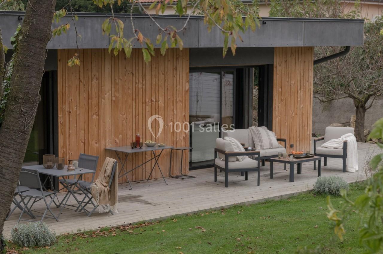 Terrasse aménagée avec mobilier de jardin devant une maison en bois, entourée de pelouse et d'arbres.