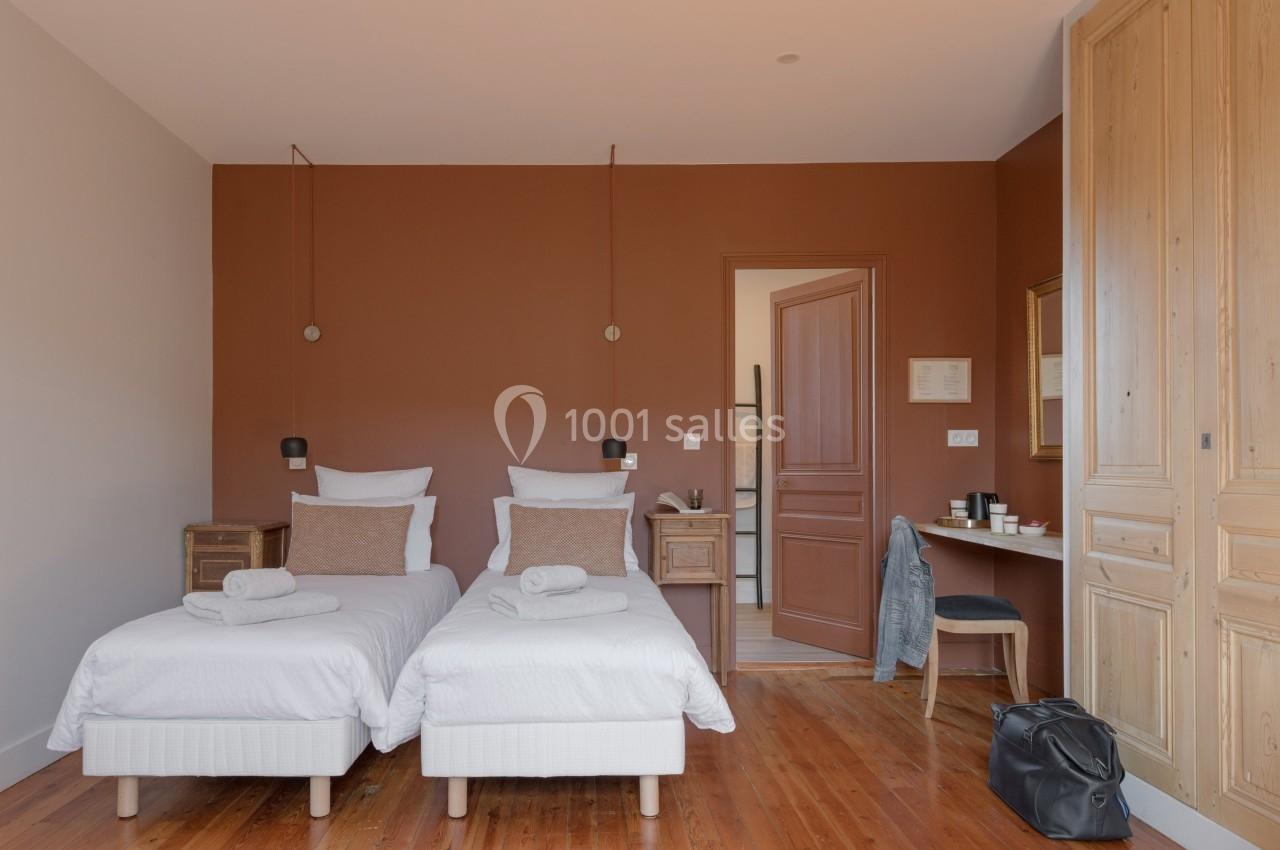 Chambre avec deux lits simples, murs marron, parquet en bois, bureau avec chaise et porte ouverte sur une autre pièce.