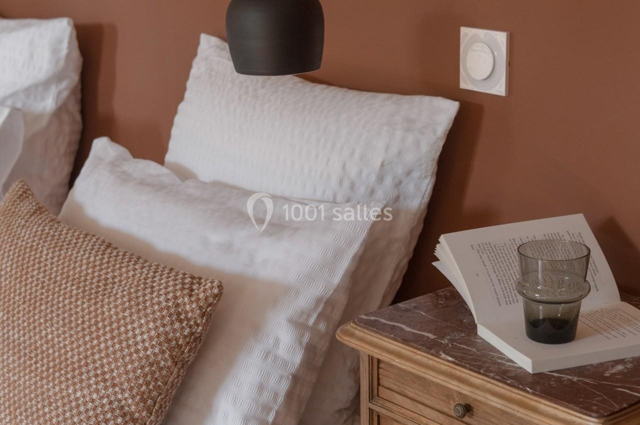 Table de chevet en bois avec un livre ouvert et un verre, à côté d'un lit aux coussins blancs et marron, mur brun en arrière…