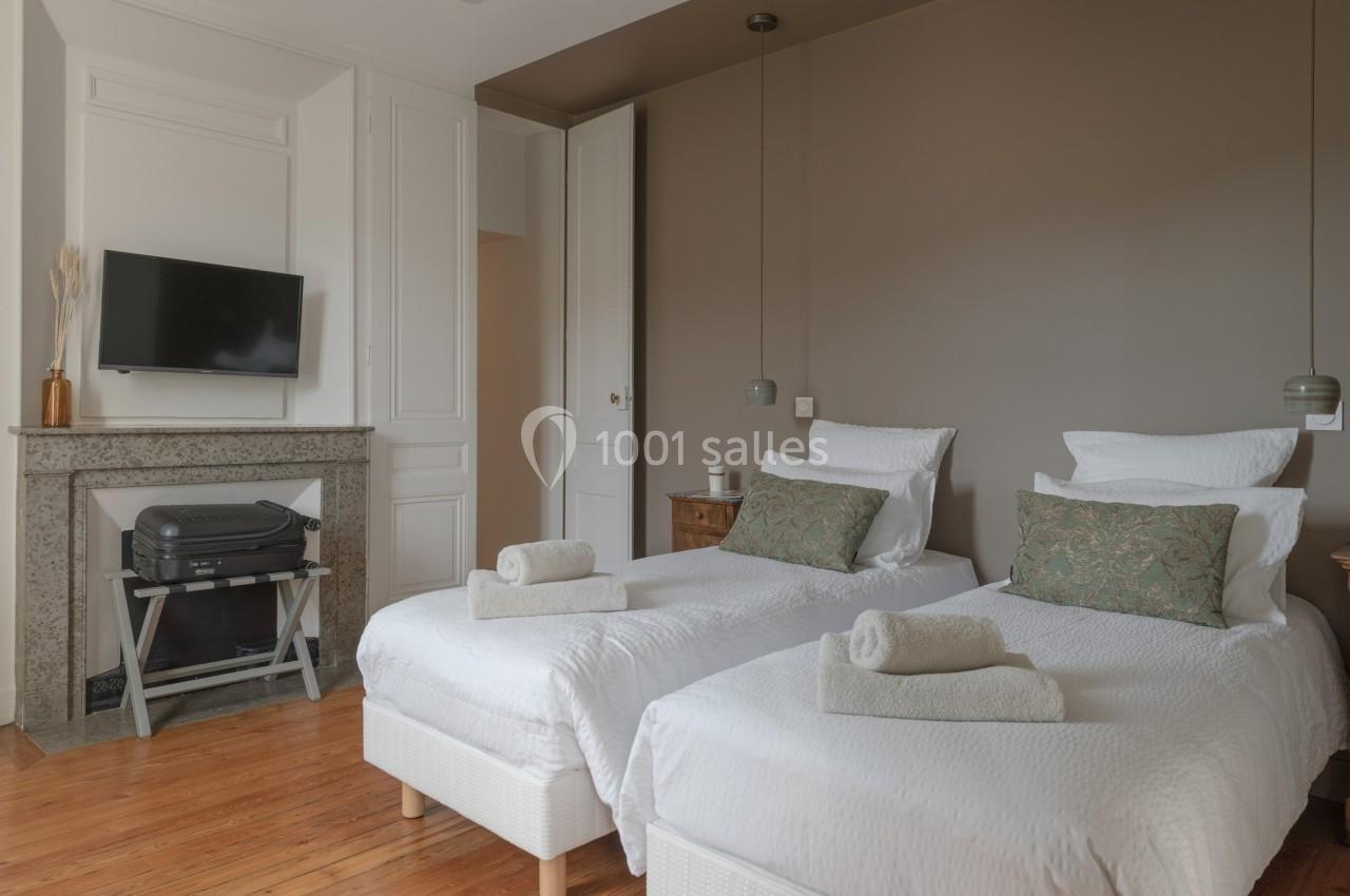 Deux lits simples avec linge blanc, coussins verts, une télévision murale et une cheminée dans une chambre lumineuse.