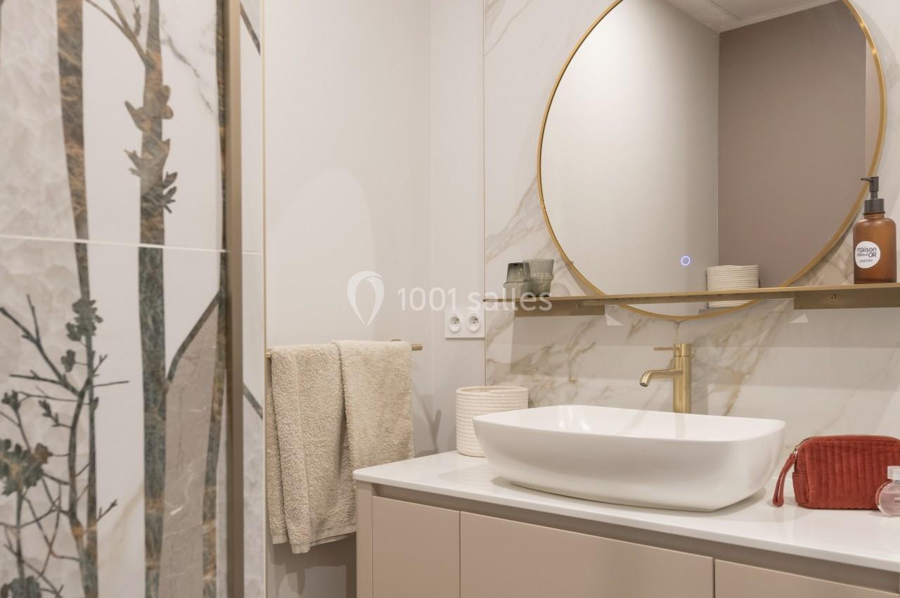 Salle de bain moderne avec lavabo blanc, miroir rond doré, carrelage effet marbre et détails décoratifs naturels.