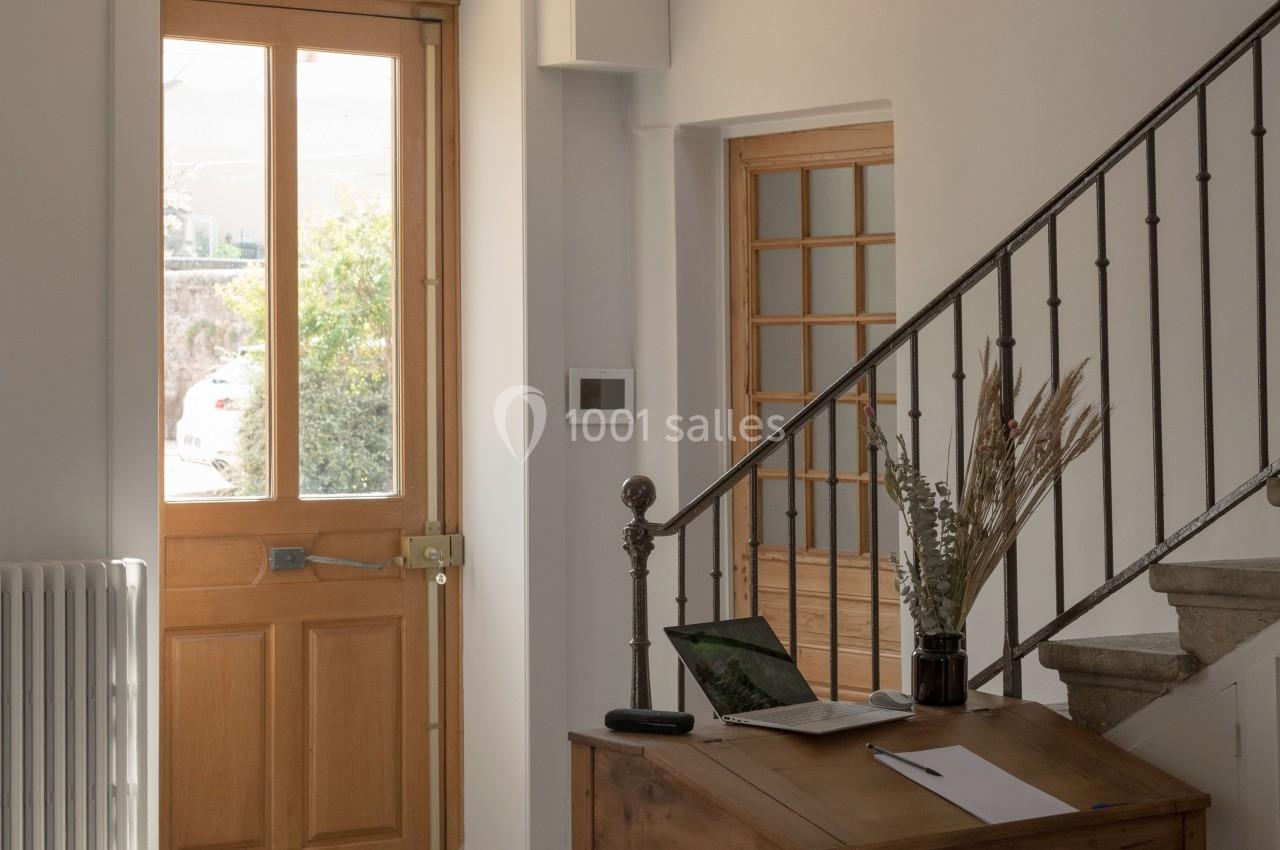 Entrée lumineuse avec porte en bois, table en bois, ordinateur portable et escalier en métal et bois.