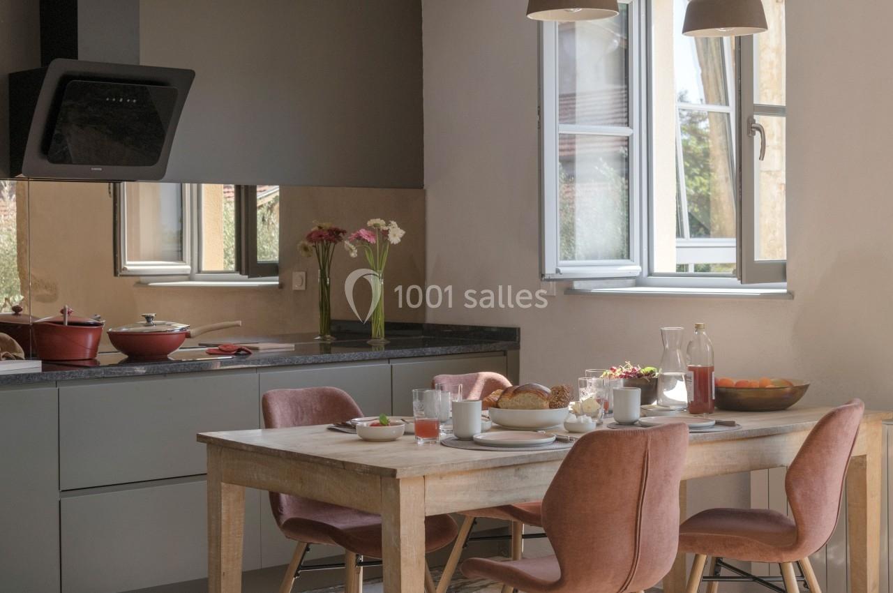 Cuisine lumineuse avec table en bois dressée pour le petit-déjeuner, chaises roses et fenêtres ouvertes sur l'extérieur.