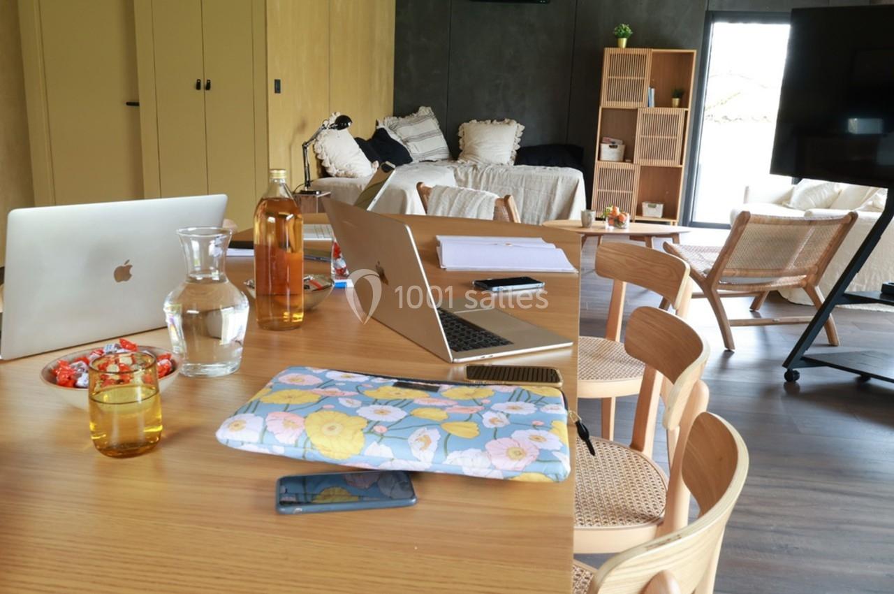 Table en bois avec ordinateurs portables, bouteille, verre d'eau et objets divers dans un salon lumineux et moderne.