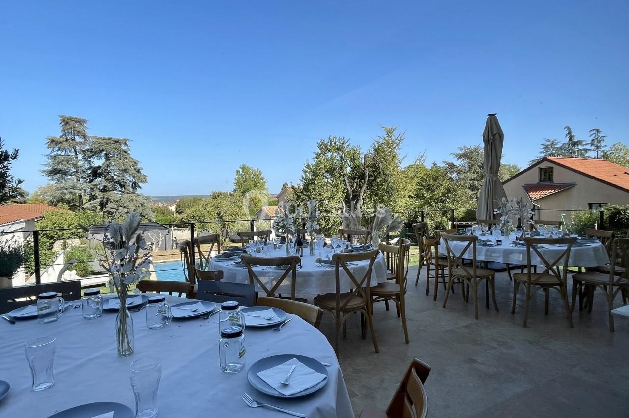 Tables rondes dressées avec vaisselle blanche sur une terrasse ensoleillée, entourée de jardins et de maisons.