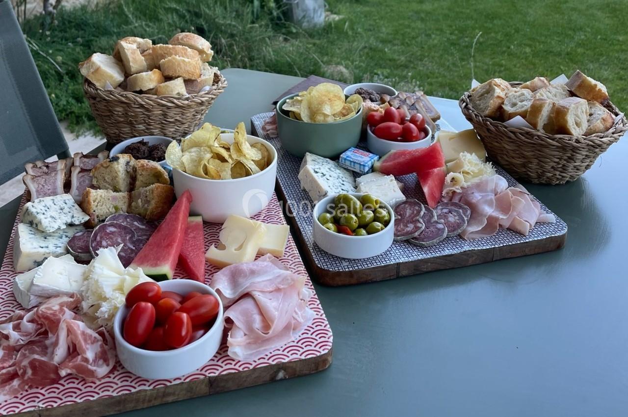 Plateaux garnis de charcuterie, fromages, fruits, olives, chips et pain, disposés sur une table en extérieur.