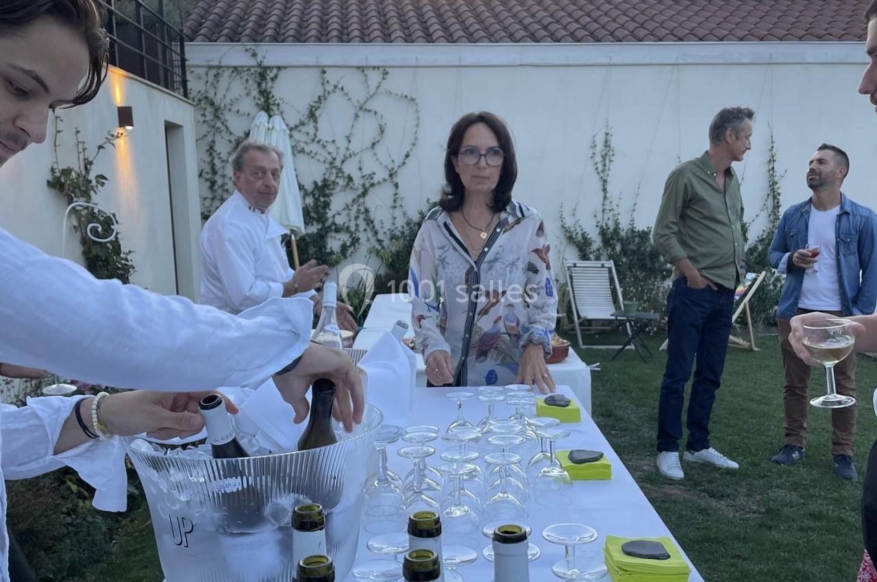 Des personnes discutent et servent des boissons autour d'une table dressée dans un jardin lors d'un événement en plein air.