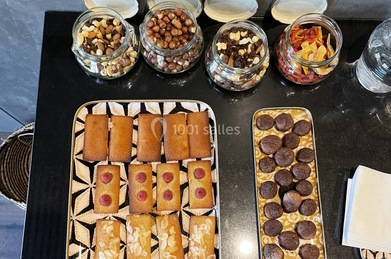 Plateaux de financiers aux amandes et framboises, biscuits au chocolat, avec pots de fruits secs en arrière-plan.