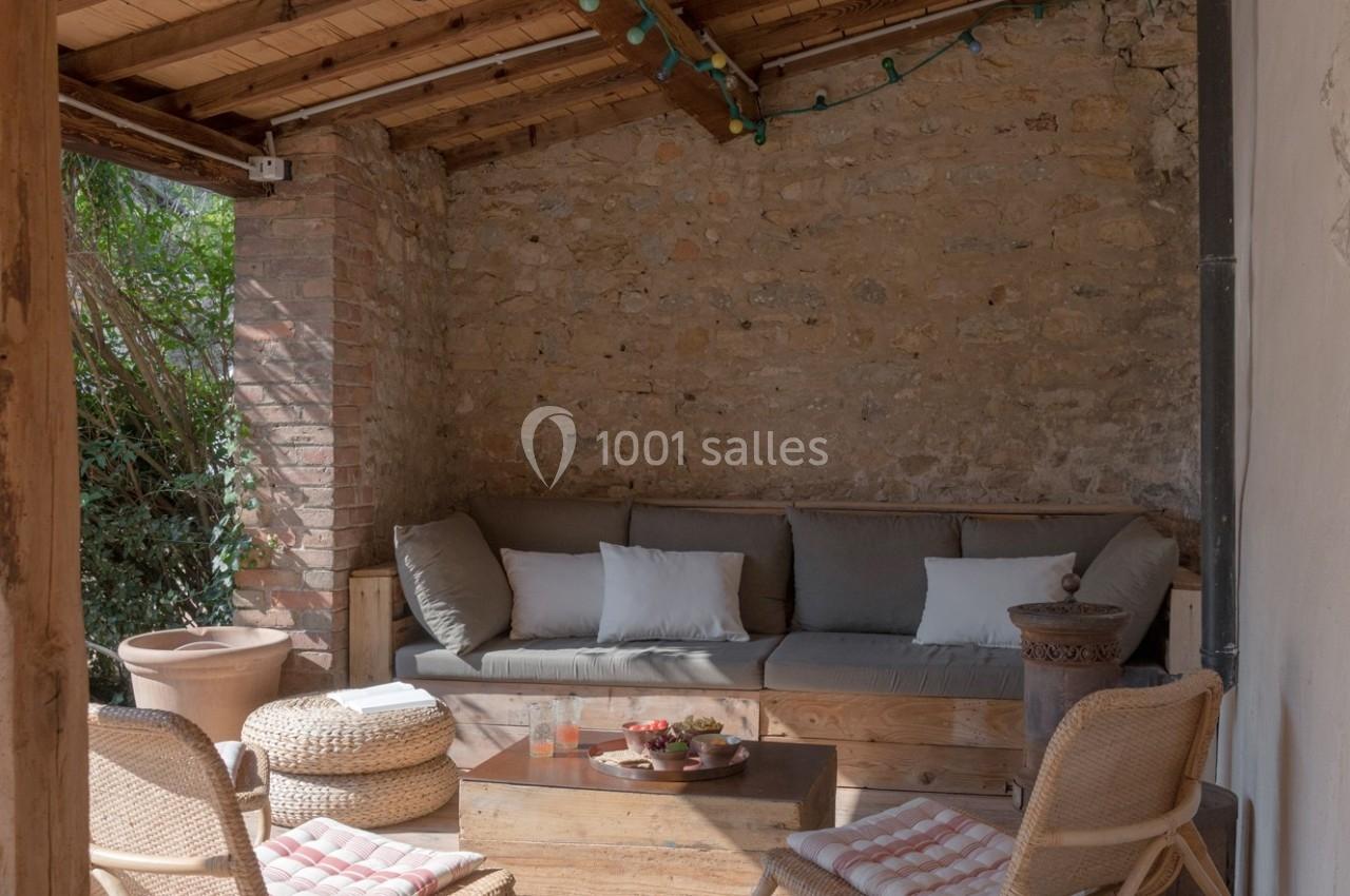 Espace salon extérieur avec canapé en bois, coussins gris et blancs, table basse et chaises en rotin sous un toit en bois.