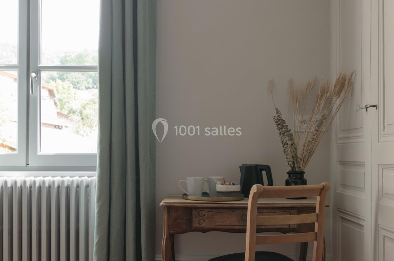 Petit bureau en bois avec une chaise, une bouilloire et une tasse, près d'une fenêtre avec rideaux verts et un radiateur.