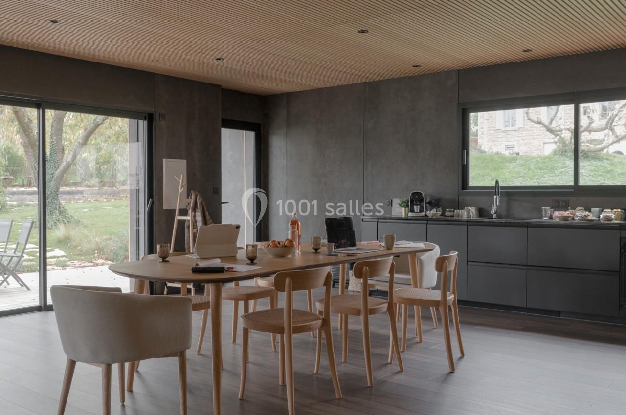 Salle à manger moderne avec table en bois, chaises assorties, cuisine ouverte et vue sur un jardin arboré.