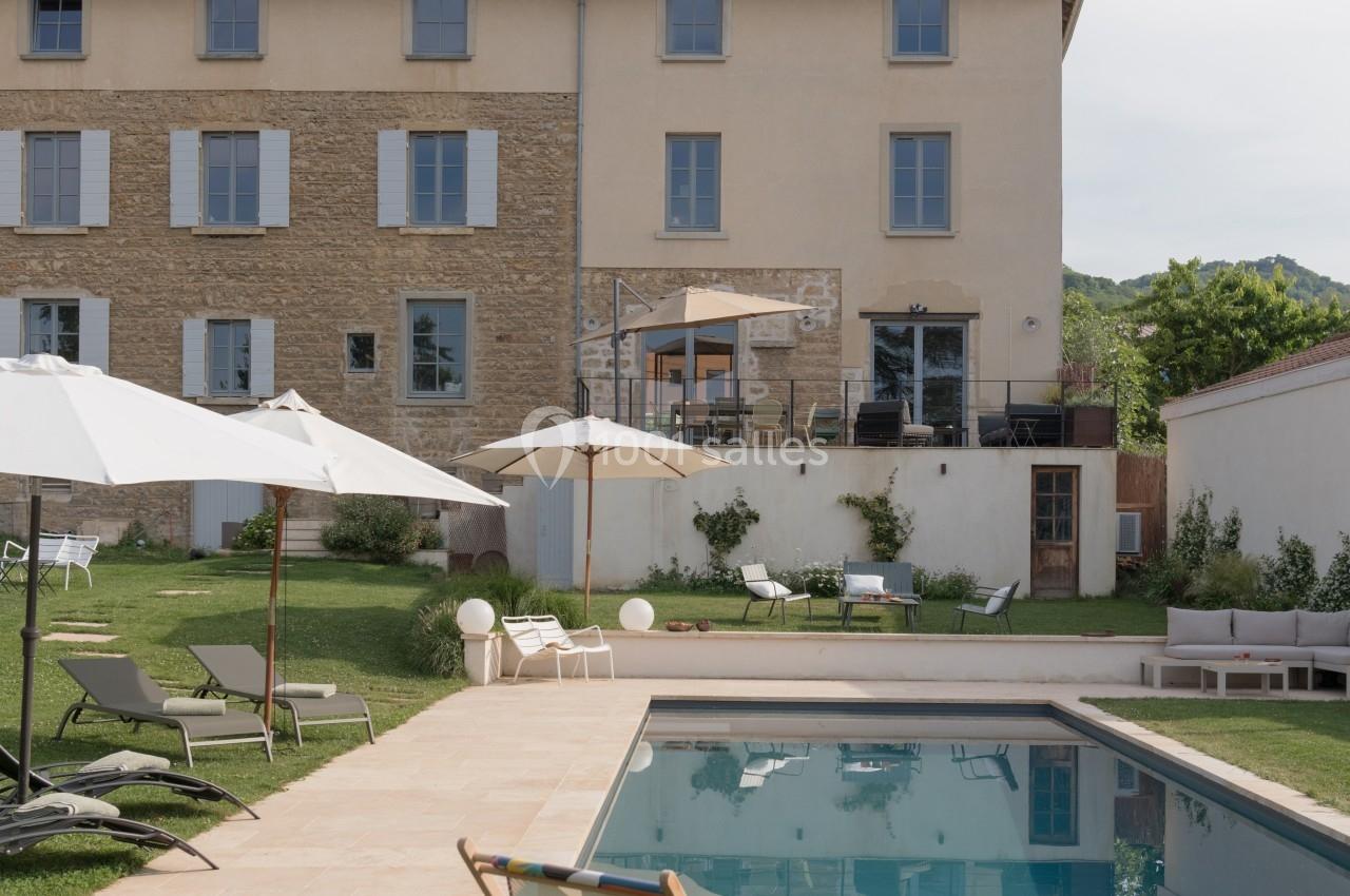 Jardin avec piscine, transats, parasols et maison en arrière-plan sous un ciel légèrement nuageux.