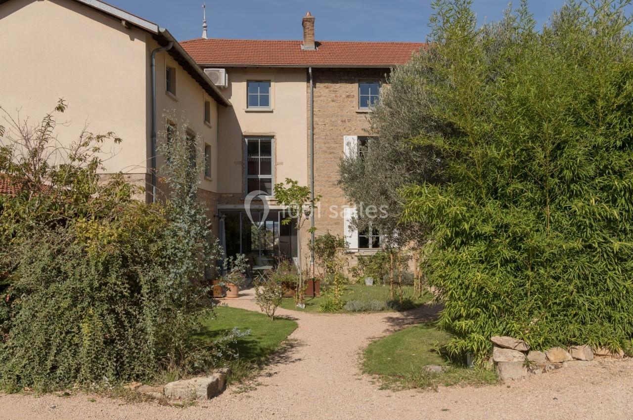 Façade d'une maison en pierre avec toit en tuiles, entourée de végétation et d'un chemin en gravier.