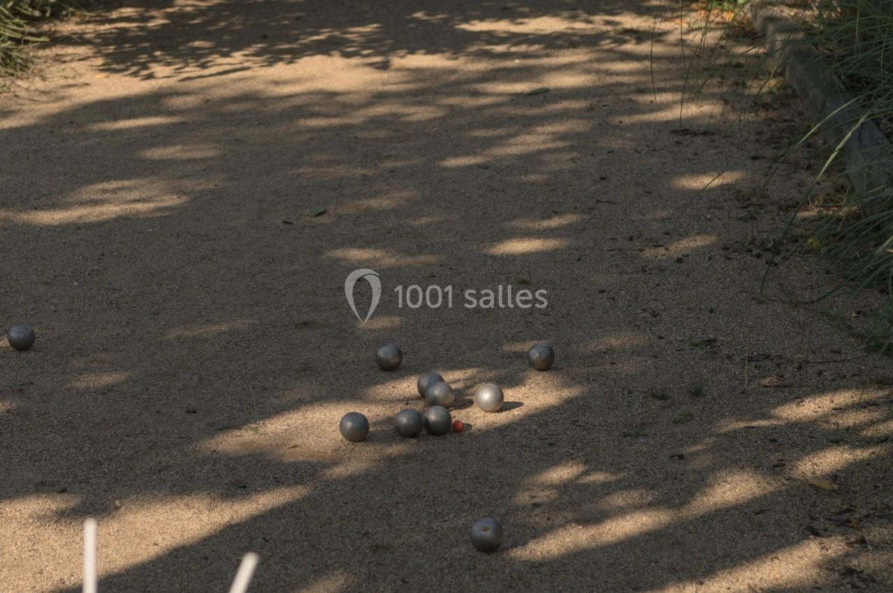 Terrain de pétanque ombragé avec des boules éparpillées au sol, deux verres de boisson posés au premier plan.